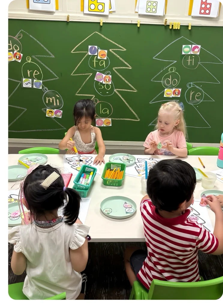Four children sitting at a table coloring worksheets in front of a chalkboard decorated with chalk-drawn Christmas trees at Creative Play Corner